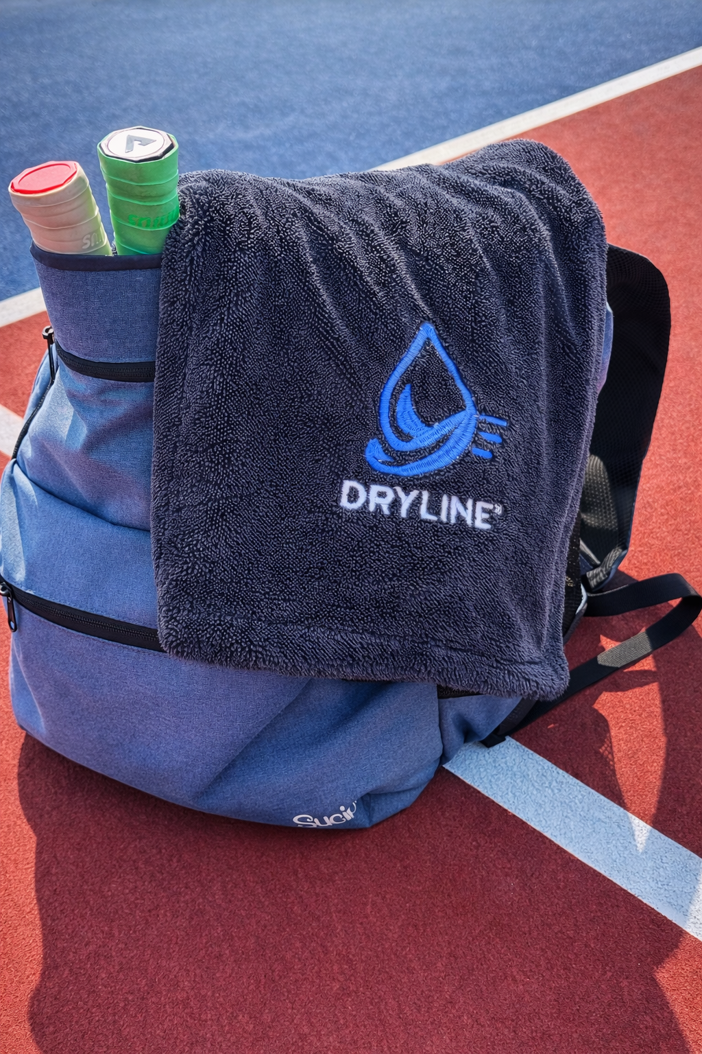 Blue backpack with towel and tennis items on a tennis court, featuring Dryline logo.